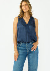 [Color: Midnight] A front facing image of a brunette model wearing a blue satin sleeveless top with an embroidered trim, elastic hem, and a surplice front. 