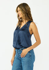 [Color: Midnight] A side facing image of a brunette model wearing a blue satin sleeveless top with an embroidered trim, elastic hem, and a surplice front. 