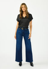 [Color: Black] A front facing image of a brunette model wearing a black satin top with an embroidered trim and short dolman sleeves. Paired with dark wash denim and black boots.