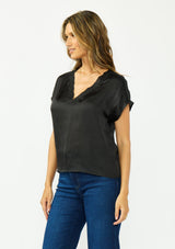 [Color: Black] A side facing image of a brunette model wearing a black satin top with an embroidered trim and short dolman sleeves. 