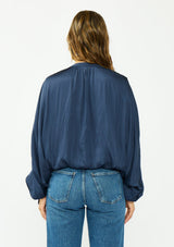 [Color: Midnight] A back facing image of a brunette model wearing a relaxed blue satin blouse with ruched sleeves and an elastic hem. 