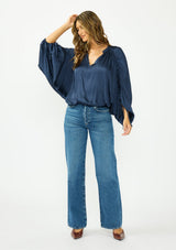 [Color: Midnight] A front facing image of a brunette model wearing a relaxed blue satin blouse with ruched sleeves and an elastic hem. 