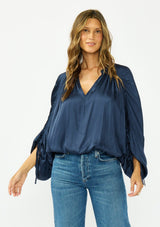 [Color: Midnight] A front facing image of a brunette model wearing a relaxed blue satin blouse with ruched sleeves and an elastic hem. 
