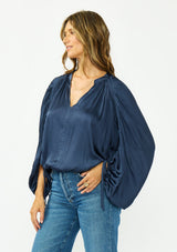 [Color: Midnight] A side facing image of a brunette model wearing a relaxed blue satin blouse with ruched sleeves and an elastic hem. 
