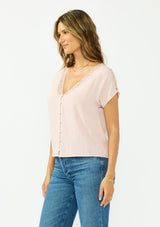 [Color: Shell] A side facing image of a brunette model wearing a pink lace trim top with short dolman sleeves and a button front. 
