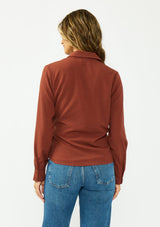 [Color: Terracotta] A back facing image of a brunette model wearing a rust button down shirt with a tie waist, collared neck, and long sleeves with button cuffs. 