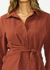 [Color: Terracotta] A detail image of a brunette model wearing a rust button down shirt with a tie waist, collared neck, and long sleeves with button cuffs. 