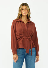 [Color: Terracotta] A front facing image of a brunette model wearing a rust button down shirt with a tie waist, collared neck, and long sleeves with button cuffs. 