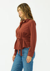 [Color: Terracotta] A side facing image of a brunette model wearing a rust button down shirt with a tie waist, collared neck, and long sleeves with button cuffs. 