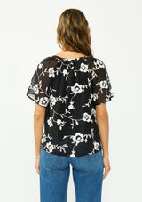 [Color: Black] A back facing image of a brunette model wearing a white and black embroidered chiffon top with short sleeves and a v neckline. 