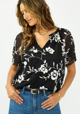 [Color: Black] A front facing image of a brunette model wearing a white and black embroidered chiffon top with short sleeves and a v neckline. 