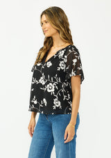 [Color: Black] A side facing image of a brunette model wearing a white and black embroidered chiffon top with short sleeves and a v neckline. 