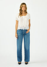 [Color: White] A front facing image of a brunette model wearing a sheer white satin top with stripes, a lace trim hem, puff sleeves, and a drawstring neckline. Styled with a dark wash denim and black heels. 