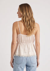[Color: Antique Blush] A back facing image of a blonde model wearing a light pink poly satin lace trim top with  a button front, adjustable spaghetti straps, and a drawstring waist with tie detail. Paired with a classic light wash denim. 