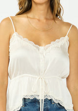[Color: Ivory] A detail image of a brunette model wearing a poly satin lace trim top with  a button front, adjustable spaghetti straps, and a drawstring waist with tie detail. Paired with a classic blue denim. 