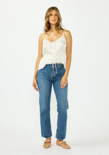 [Color: Ivory] A front facing image of a brunette model wearing a poly satin lace trim top with  a button front, adjustable spaghetti straps, and a drawstring waist with tie detail. Paired with a classic blue denim and nude ballet flats.