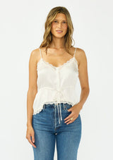 [Color: Ivory] A front facing image of a brunette model wearing a poly satin lace trim top with  a button front, adjustable spaghetti straps, and a drawstring waist with tie detail. Paired with a classic blue denim. 