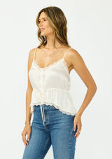 [Color: Ivory] A side facing image of a brunette model wearing a poly satin lace trim top with  a button front, adjustable spaghetti straps, and a drawstring waist with tie detail. Paired with a classic blue denim. 
