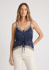 [Color: Navy/Taupe] A front facing image of a blonde model wearing a navy blue poly satin lace trim top with  a button front, adjustable spaghetti straps, and a drawstring waist with tie detail. Paired with a classic off white denim. 