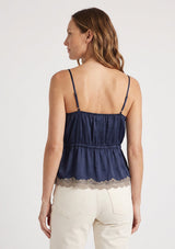 [Color: Navy/Taupe] A back facing image of a blonde model wearing a navy blue poly satin lace trim top with  a button front, adjustable spaghetti straps, and a drawstring waist with tie detail. Paired with a classic off white denim. 