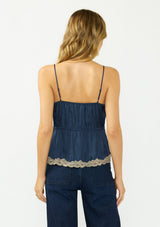 [Color: Navy/Taupe] A back facing image of a blonde model wearing a navy blue poly satin lace trim top with  a button front, adjustable spaghetti straps, and a drawstring waist with tie detail. Paired with a classic dark wash denim.