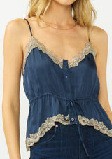 [Color: Navy/Taupe] A detail image of a blonde model wearing a navy blue poly satin lace trim top with  a button front, adjustable spaghetti straps, and a drawstring waist with tie detail. Paired with a classic dark wash denim.