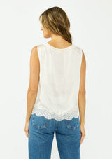 [Color: Ivory] A back facing image of a brunette model wearing an ivory poly satin tank top with a ruffled v neckline, tie front, button front, and a lace trim hem. 