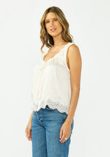 [Color: Ivory] A side facing image of a brunette model wearing an ivory poly satin tank top with a ruffled v neckline, tie front, button front, and a lace trim hem. 