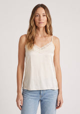 [Color: Champagne] A front facing image of a blonde model wearing an ivory satin camisole with lace trim, adjustable spaghetti straps, and side slits. 