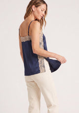 [Color: Navy/Taupe] A side facing image of a blonde model wearing a navy satin camisole with lace trim, adjustable spaghetti straps, and side slits. 