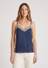 [Color: Navy/Taupe] A front facing image of a blonde model wearing a navy satin camisole with lace trim, adjustable spaghetti straps, and side slits. 