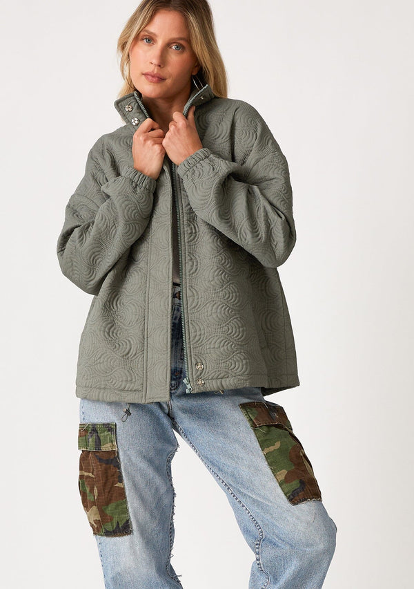 [Color: Olive] A front facing image of a blonde model wearing an olive green quilted jacket. With a zip front, long sleeves, a drawstring waist with adjustable toggles, and a funnel neck.