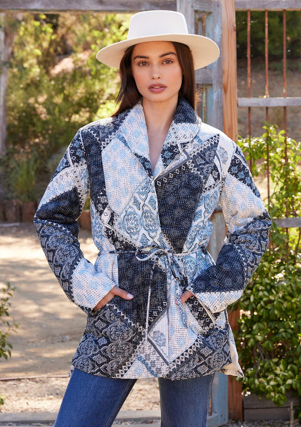 [Color: Natural/Blue] A front facing image of a brunette model standing outside wearing a blue and white patchwork print quilted jacket. With long sleeves, a shawl collar, side pockets, and a tie waist belt.
