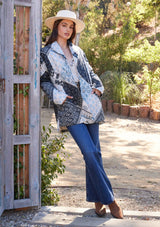 [Color: Natural/Blue] A full body front facing image of a brunette model standing outside wearing a blue and white patchwork print quilted jacket. With long sleeves, a shawl collar, side pockets, and a tie waist belt.