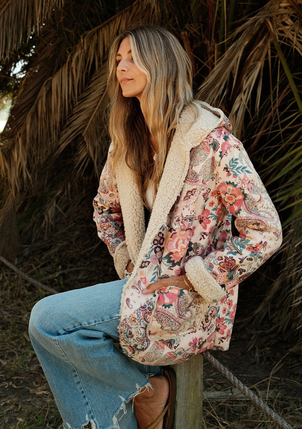 [Color: Natural/Red] A side facing image of a blonde model wearing a bohemian jacket with a pink and natural floral print and a faux shearling lining. With long sleeves, side pockets, a hoodie, and an open front.