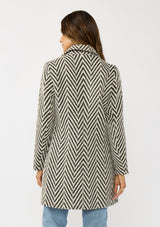 [Color: Cream/Black] A back facing image of a brunette model wearing a classic women's coat in a cream and black chevron stripe. With long sleeves, a notched collar, a button front, and side pockets. 