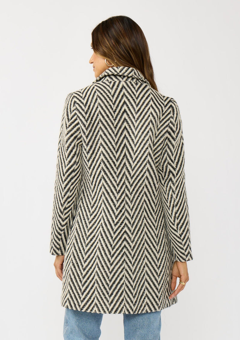 [Color: Cream/Black] A back facing image of a brunette model wearing a classic women's coat in a cream and black chevron stripe. With long sleeves, a notched collar, a button front, and side pockets. 
