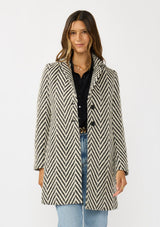 [Color: Cream/Black] A front facing image of a brunette model wearing a classic women's coat in a cream and black chevron stripe. With long sleeves, a notched collar, a button front, and side pockets. 