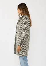 [Color: Cream/Black] A side facing image of a brunette model wearing a classic women's coat in a cream and black chevron stripe. With long sleeves, a notched collar, a button front, and side pockets. 