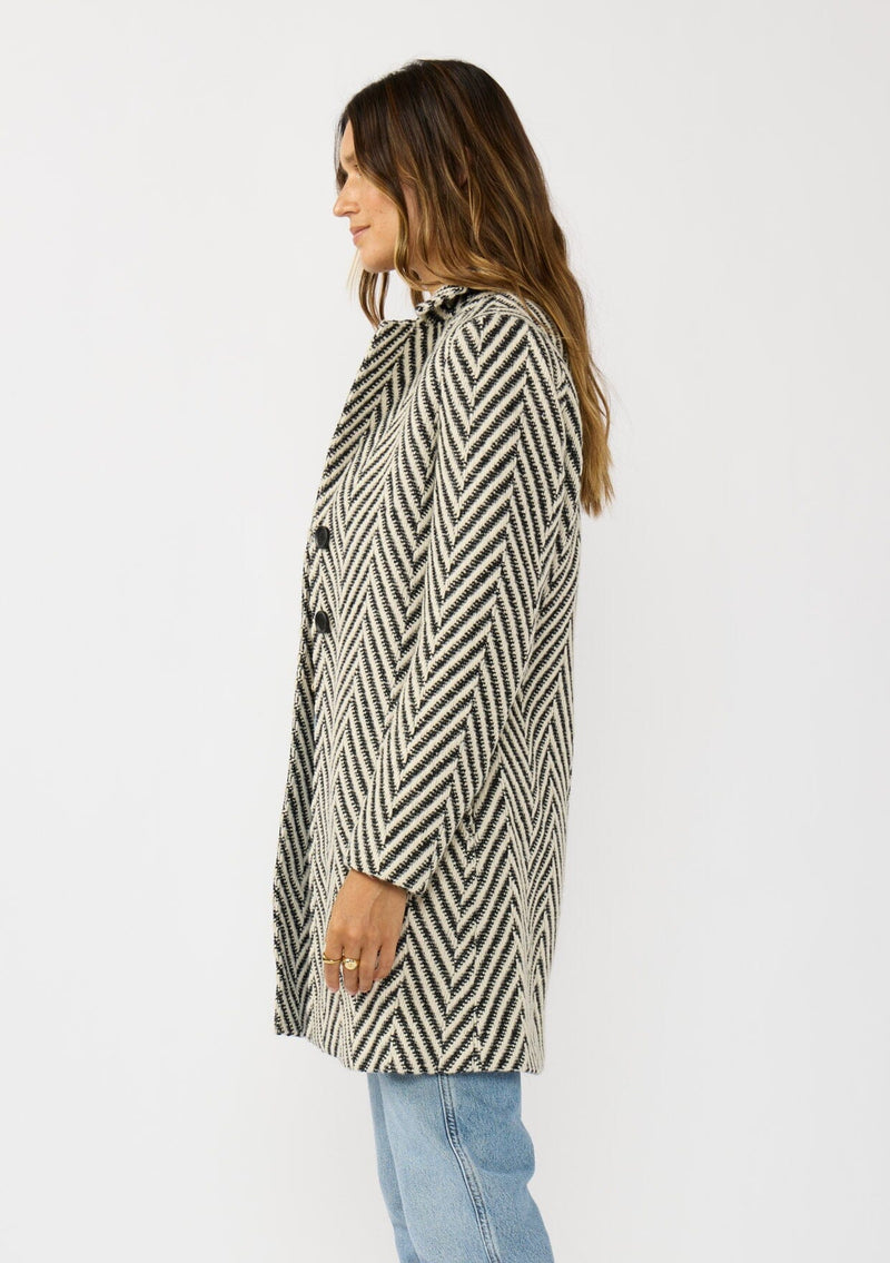 [Color: Cream/Black] A side facing image of a brunette model wearing a classic women's coat in a cream and black chevron stripe. With long sleeves, a notched collar, a button front, and side pockets. 