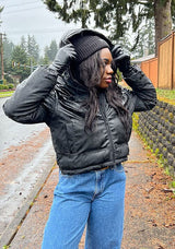 [Color: Black] A half body front facing image of a black woman wearing an ultra puffy cropped jacket in a matte black finish. Featuring an adjustable hoodie with toggles, a zippered front, and side pockets.