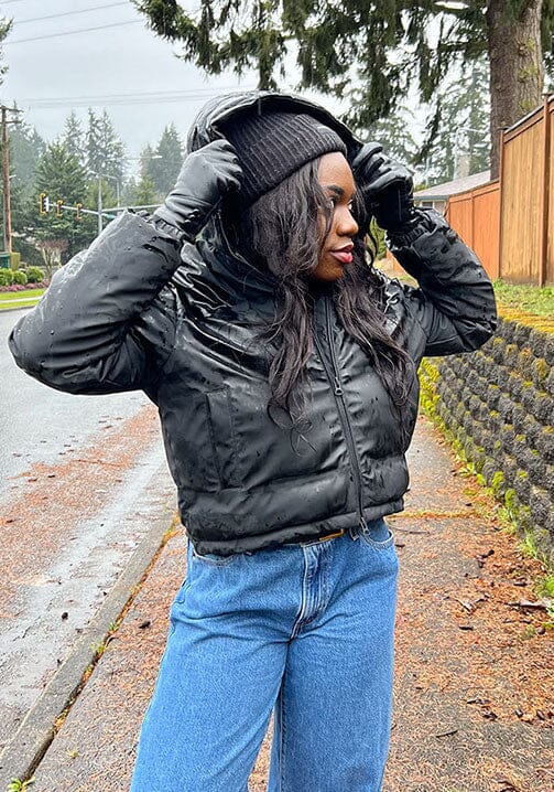 [Color: Black] A half body front facing image of a black woman wearing an ultra puffy cropped jacket in a matte black finish. Featuring an adjustable hoodie with toggles, a zippered front, and side pockets.