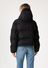 [Color: Black] A back facing image of a blonde model wearing an ultra puffy cropped jacket in a matte black finish. Featuring an adjustable hoodie with toggles, a zippered front, and side pockets.