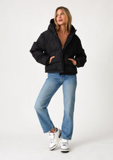 [Color: Black] A full body front facing image of a blonde model wearing an ultra puffy cropped jacket in a matte black finish. Featuring an adjustable hoodie with toggles, a zippered front, and side pockets.