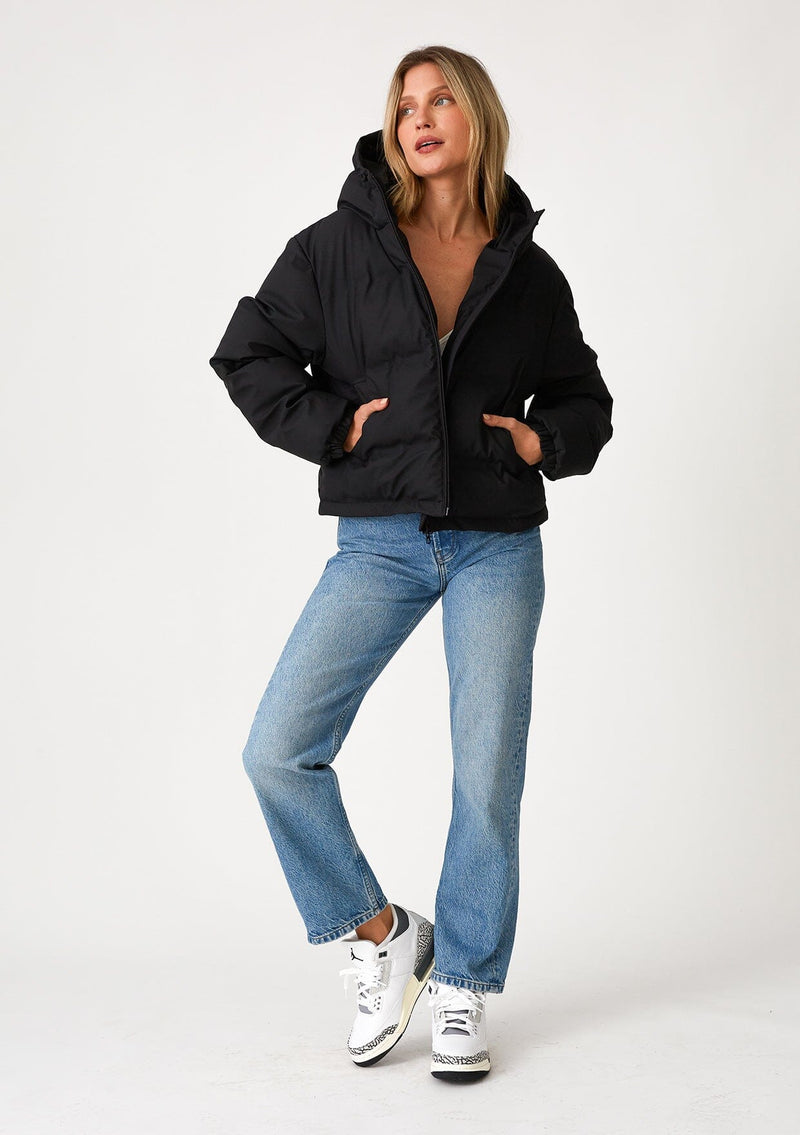 [Color: Black] A full body front facing image of a blonde model wearing an ultra puffy cropped jacket in a matte black finish. Featuring an adjustable hoodie with toggles, a zippered front, and side pockets.