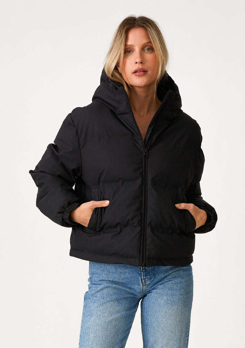 [Color: Black] A front facing image of a blonde model wearing an ultra puffy cropped jacket in a matte black finish. Featuring an adjustable hoodie with toggles, a zippered front, and side pockets.
