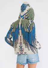 [Color: Natural/Teal] A back facing image of a brunette model wearing a bohemian statement hoodie in a natural and teal blue peacock tapestry design. With long sleeves, a front zip closure, side pockets, and a drawstring hoodie.
