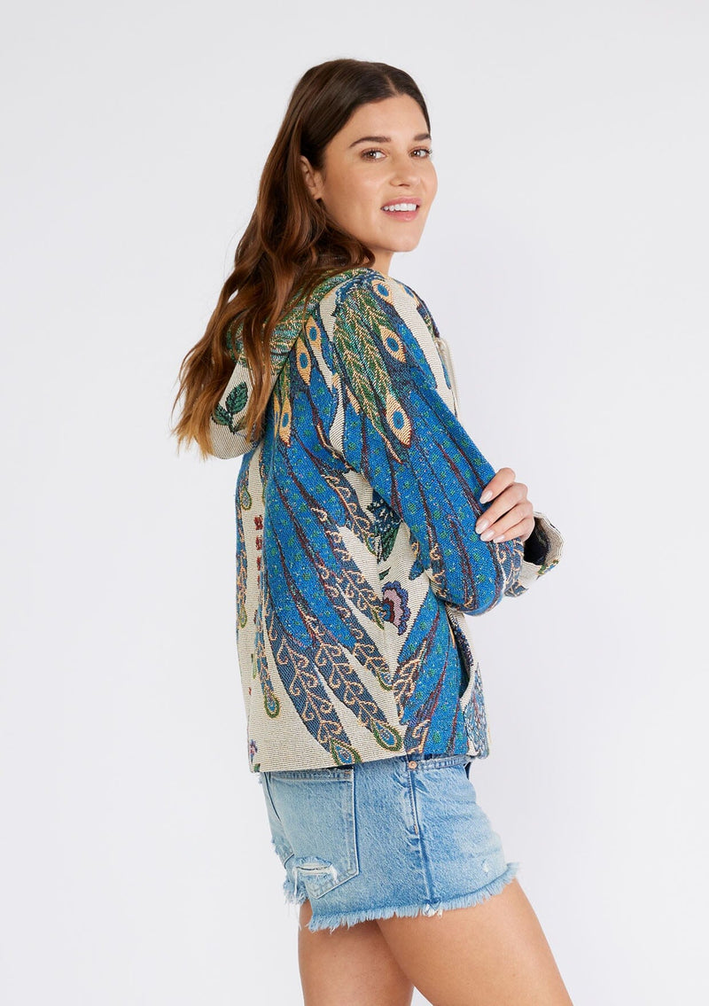 [Color: Natural/Teal] A side facing image of a brunette model wearing a bohemian statement hoodie in a natural and teal blue peacock tapestry design. With long sleeves, a front zip closure, side pockets, and a drawstring hoodie. 