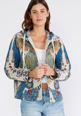 [Color: Natural/Teal] A front facing image of a brunette model wearing a bohemian statement hoodie in a natural and teal blue peacock tapestry design. With long sleeves, a front zip closure, side pockets, and a drawstring hoodie.