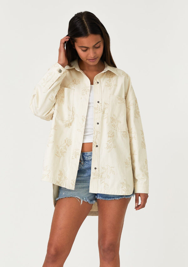 [Color: Natural] A front facing image of a brunette model wearing an off white bohemian shirt jacket crafted from cotton. With long sleeves, a collared neckline, a snap button front, front patch pockets, and tonal embroidered detail throughout. 